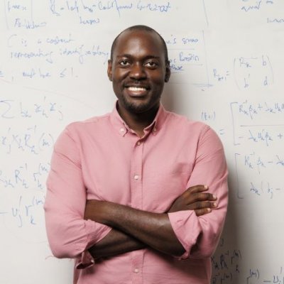 A man in a pink shirt stands smiling broadly with his arms crossed in front of a whiteboard covered in mathematical equations. 